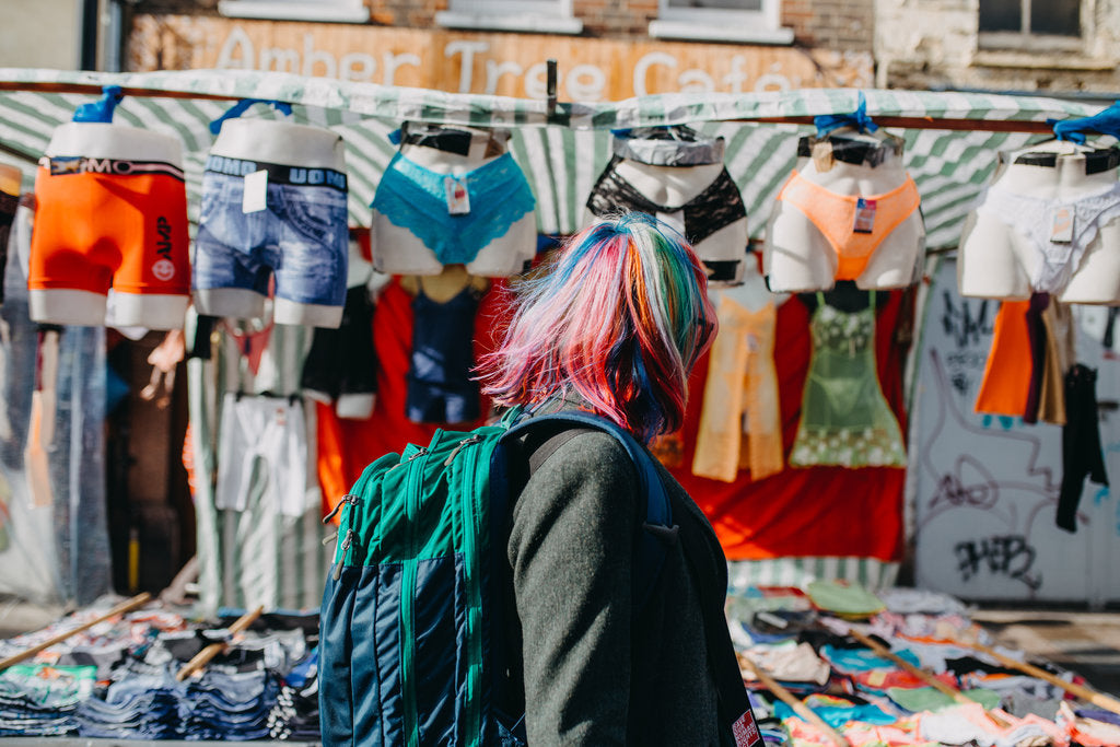 The author with rainbow hair, walking through Deptford Market. The background features an array of multicoloured pants on mannequins.